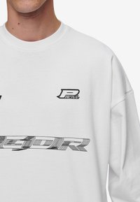 White sweatshirt with black graphic text and logos. Round neckline, loose fit, soft fabric. Features "P" logo and stylized lettering.
