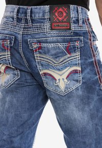Blaue Jeans aus Denim mit bestickten Akzenten, versehen mit einem dunklen Patch-Label und dekorativen Nähten um die Gesäßtaschen.