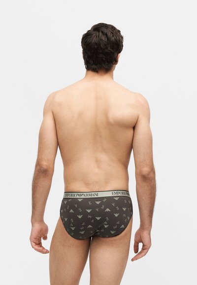 Άνδρας μοντέλο που στέκεται με την πλάτη προς τα μπρος, φοράει σκούρα εσώρουχα Emporio Armani με μοτίβο λογότυπου σε απλό ανοιχτό φόντο.