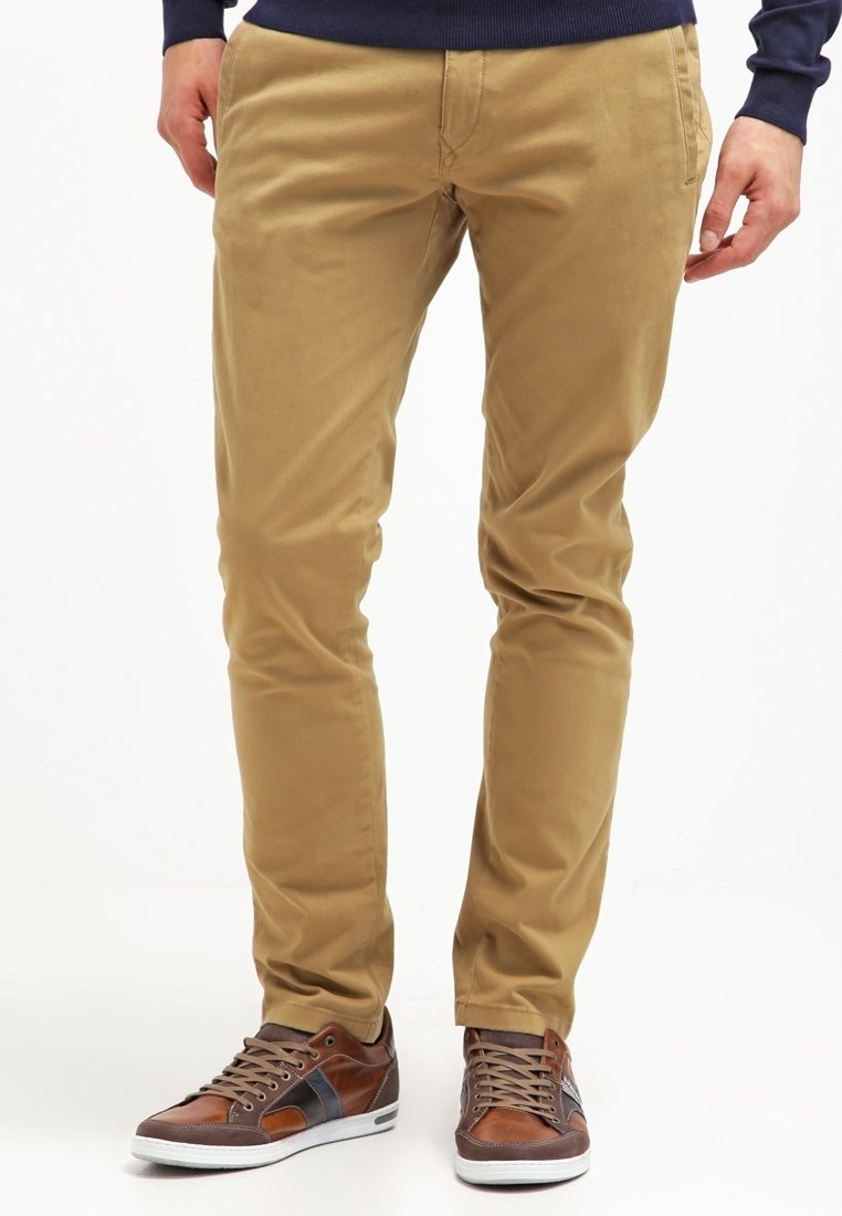 Homme portant un pantalon beige slim et des baskets en cuir marron avec des accents foncés, debout devant un fond blanc.