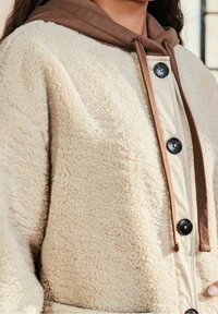 Maglione in pile color crema con cappuccio marrone. Presenta grandi bottoni neri e un pannello frontale beige a contrasto. Texture morbida e design sportivo.