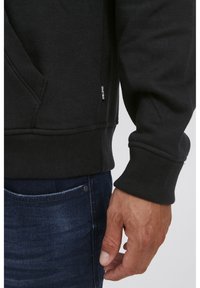 Sudadera negra hecha de mezcla de algodón; presenta puños y dobladillo acanalados, bolsillo frontal y una etiqueta de marca sutil en el lateral.