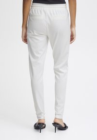White tapered trousers with elastic waistband and two back pockets, styled with black high-heeled shoes. Smooth texture, minimalist design.