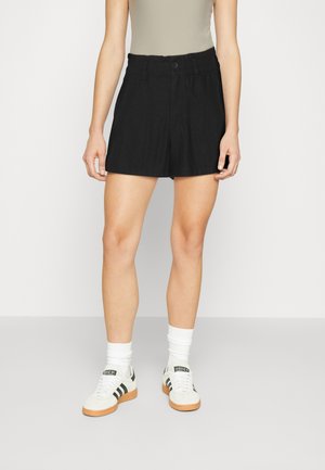 Pantalones cortos negros de corte relajado, con cinturilla elástica y cierre de botón. Combinados con calcetines blancos y zapatillas a rayas negras.