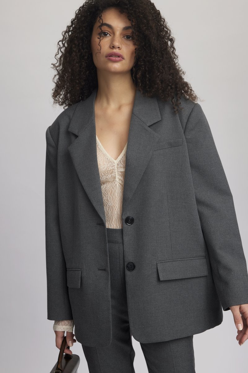 Sivý oversized blazer z textúrovanej látky, s klopami, dvoma gombíkmi a prednými vreckami, nosený cez čipkový top.
