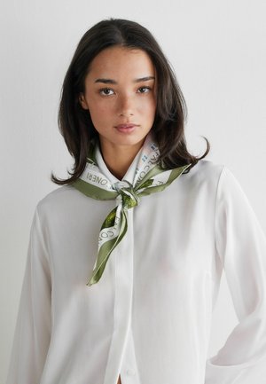 Jeune femme aux cheveux foncés mi-longs porte une chemise blanche boutonnée et une écharpe à motifs blanc et vert nouée autour du cou, fond neutre.