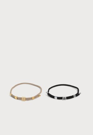 To tynde reb-armbånd med metal accenter, en beige med gulddetaljer og en sort med sølvdetaljer, hver med en firkantet logo charm.