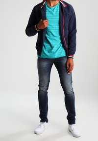 Marinblå kavaj med röda och vita detaljer, turkosrandig t-shirt, blå jeans och vita sneakers. Avslappnad, lager-på-lager-outfit.