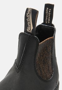 Bottines Chelsea en cuir noir avec des panneaux élastiques sur les côtés, mettant en valeur un subtil accent métallique. Détails du logo et de la marque sur les languettes.