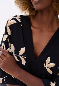 Zwarte kleding met een diepe V-hals en losse mouwen, voorzien van een crèmekleurig bladpatroon. Gelaagd stof met een zachte val en natuurlijke beweging.