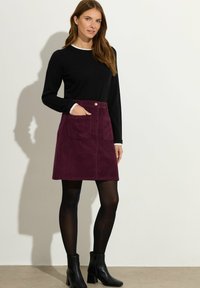 Femme portant un haut noir à manches longues, une jupe en velours côtelé violet avec des poches, des collants noirs et des bottines noires, se tenant devant un fond uni.