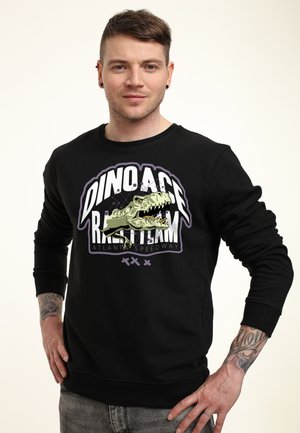 Young man with short brown hair wearing a black sweatshirt featuring a dinosaur head and "DINO ACE RACING TEAM" text.