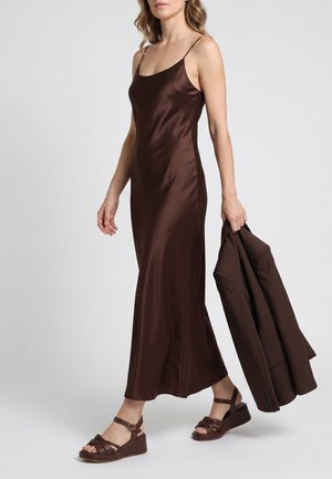 Kvinde iført en brun satin natkjole og matchende sandaler, der holder en brun blazer i venstre hånd, stående mod en ensartet lys baggrund.