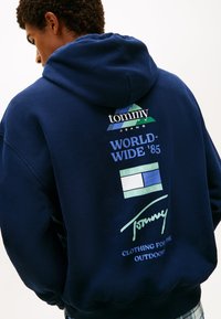 Μπλε σκούρο φούτερ με κουκούλα, με λογότυπο και κείμενο στην πλάτη. Διαθέτει γεωμετρικά σχέδια σε πράσινο και μπλε, με άνετη εφαρμογή και υφή υφάσματος.