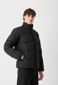 Jachetă puffer neagră cu guler înalt, design matlasat și închidere cu fermoar. Dispune de un buzunar lateral și un accent cu logo la tiv.