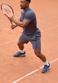 Hall valge sokid ja mustad tenniseketid, mängija valmistub lööma tennisepalli punase reketiga savikattega väljakul.