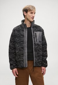 Čierna a šedá vzorovaná fleecová bunda s vysokým golierom, zipsom na hrudníku a kontrastnými šedými rukávmi. Nosená na čiernej košeli.