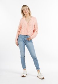 Cardigan lavorato a maglia rosa chiaro con scollo a V, caratterizzato da un motivo a treccia e bottoni di dimensioni medie. Abbinato a jeans azzurri e sneakers bianche.