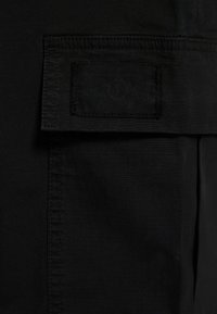 Svart tyg med en texturerad yta; har en ficka med ett kardborreband. Rak sömdetalj synlig längs kanterna.