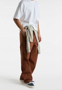 Hombre con camiseta blanca de gran tamaño, pantalones sueltos marrones, sudadera gris atada a la cintura y zapatillas azul-grises sobre fondo blanco.