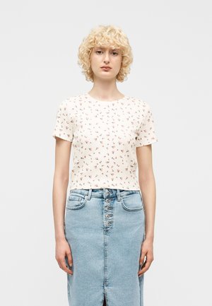 Kvinne med krøllete blondt hår iført en hvit kortermet blomstret topp og lys blå denimskjørt med høyt liv og knapper foran.