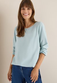 Helle blaue Bluse aus glattem Stoff, runder Ausschnitt mit einer kleinen Knopfdetails, lange Ärmel leicht hochgerollt, kombiniert mit dunklen Jeans.