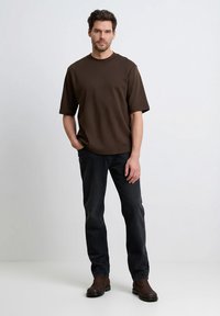 Brązowy t-shirt z krótkim rękawem i okrągłym dekoltem, w połączeniu z ciemnymi dżinsami i brązowymi butami, stojący na jasnym tle. Prosty krój i design.