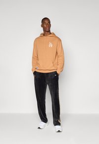 Hoodie marron avec logo blanc, poche avant et cordon de serrage. Pantalons en velours noir avec bandes latérales et poches. Baskets blanches.