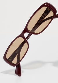 Óculos de sol bordeaux com armações quadradas, lentes com tonalidade castanha e laterais texturizadas. Apresenta o logótipo na haste e um acabamento liso e brilhante.