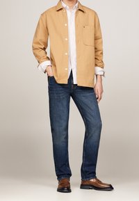 Khaki Baumwollhemd über einem weiß gestreiften Hemd, kombiniert mit dunkelblauen Jeans und braunen Schuhen; glatte Textur, ansprechendes Design, offener Kragen.