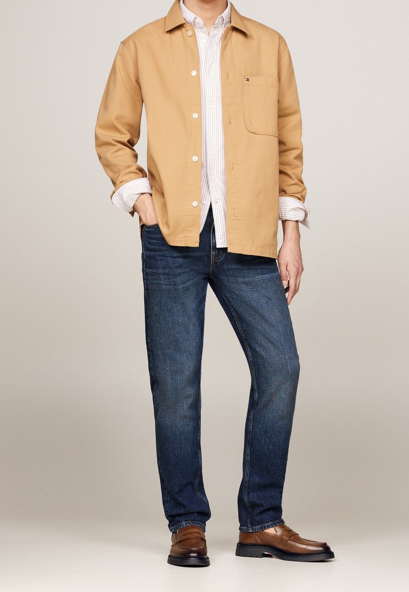 Khaki Baumwollhemd über einem weiß gestreiften Hemd, kombiniert mit dunkelblauen Jeans und braunen Schuhen; glatte Textur, ansprechendes Design, offener Kragen.