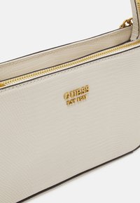 Beige, gestructureerde synthetische handtas met goudkleurige metalen accenten, ritssluiting en reliëflogo. Heeft een slank, elegant ontwerp.