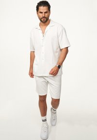 Weißes kurzärmliges Hemd und dazu passende Shorts, die beide ein strukturiertes Muster aufweisen. Kombiniert mit weißen Sneakern und Socken mit schwarzen Streifen.