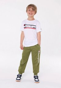 Niño sonriente de pie con las manos en los bolsillos, vistiendo una camiseta blanca de Champion, pantalones de chándal verdes de Champion y zapatillas negras y blancas.