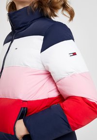 Padded jacket featuring a high collar, color-blocked design in navy, white, pink, and red, with zip closure and logo detail on the sleeve.