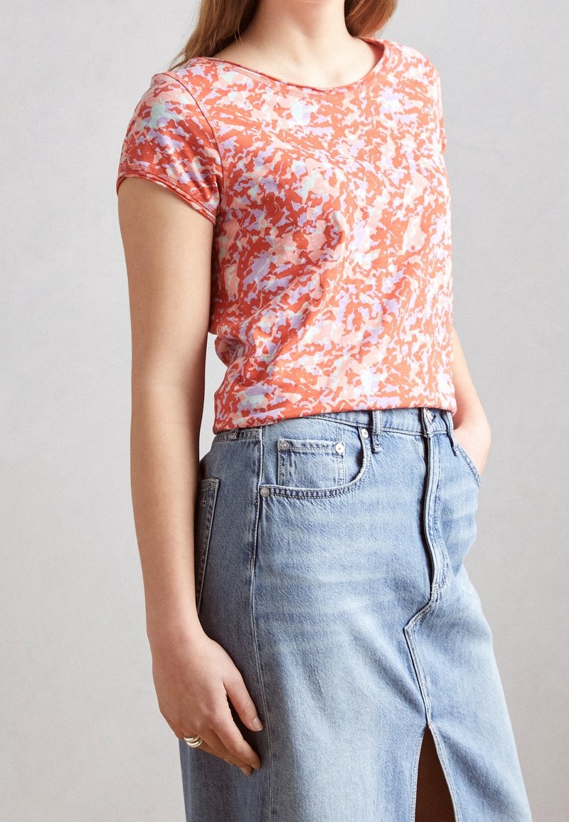 Femme portant un haut à manches courtes rouge et violet clair à motifs, rentré dans une jupe en jean bleu clair avec une fente à l'avant.