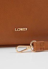 Borsa in pelle marrone con texture, dotata di un logo dorato "L.CREDI" e di un attacco in metallo dorato su una tracolla coordinata.