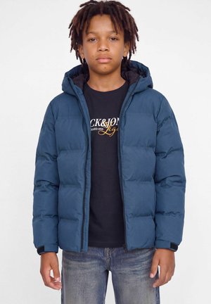 Un garçon avec des petites dreadlocks courtes se tient devant un fond blanc, portant une doudoune bleue à capuche, un T-shirt noir avec un motif graphique et un jean.