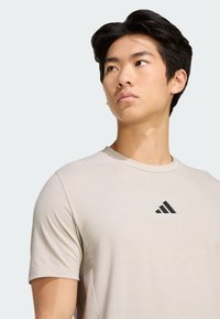 Jasno beżowa koszulka sportowa wykonana z miękkiego materiału, z czarnym logo Adidasa na lewej piersi i krótkimi, zaokrąglonymi rękawami.