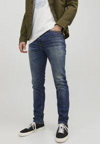 Jeans skinny in denim blu con dettagli sbiaditi, abbinati a una t-shirt bianca e a una giacca verde oliva. Sneakers nere con lacci bianchi.