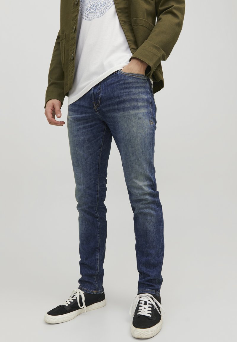 Jeans skinny in denim blu con dettagli sbiaditi, abbinati a una t-shirt bianca e a una giacca verde oliva. Sneakers nere con lacci bianchi.