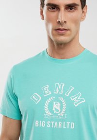 Tirkīza kokvilnas T-kreklam ir apaļš kakls un balta grafikas druka ar tekstu "DENIM" un vainaga dizainu, marķēts ar "BIG STAR LTD."