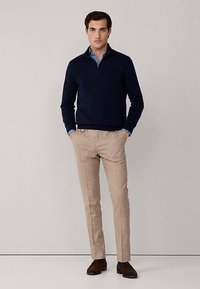 Námořnický pulovr se zipovým límcem v kombinaci se světlými béžovými kalhotami. Hnědé boty dokončují vzhled, který se vyznačuje upraveným střihem a hladkou texturou.