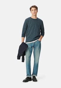 Marineblauwe gebreide sweatshirt over een witte shirt, gecombineerd met lichtblauwe jeans, zwarte schoenen en een marineblauwe trui over de arm geslagen.