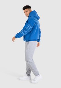 Blauwe hoodie met witte letters op de mouwen, gecombineerd met grijze sweatpants. Model draagt witte sneakers. Grote, soepele stof, relaxte pasvorm.