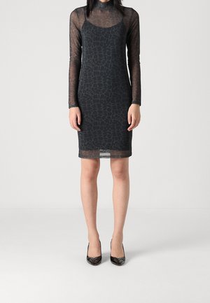 Frau trägt ein schwarzes, durchsichtiges, langärmliges Kleid mit Muster und Unterkleid sowie schwarze spitze High Heels, steht vor neutralem Hintergrund.