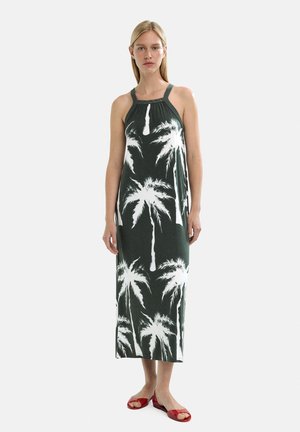 Maxi sukienka w ciemnozielonym kolorze z białym nadrukiem palm; projekt z dekoltem na halter z małymi okrągłymi detalami. Modelka ubrana w czerwone płaskie sandały.
