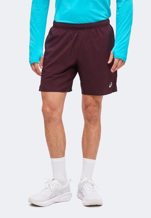 Homme portant un short de sport bordeaux, des chaussettes blanches, des chaussures de course blanches et un haut turquoise à manches longues, debout devant un fond blanc.
