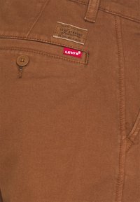 Jean en denim marron avec une poche avant, comportant une étiquette de marque et une étiquette rouge. La texture est lisse avec des détails de couture visibles.