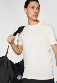 Μπεζ βαμβακερό t-shirt με κεντημένο λογότυπο «Champion». Κοντά μανίκια, στρογγυλή λαιμόκοψη, λεία υφή. Μαύρη αθλητική τσάντα στο φόντο.